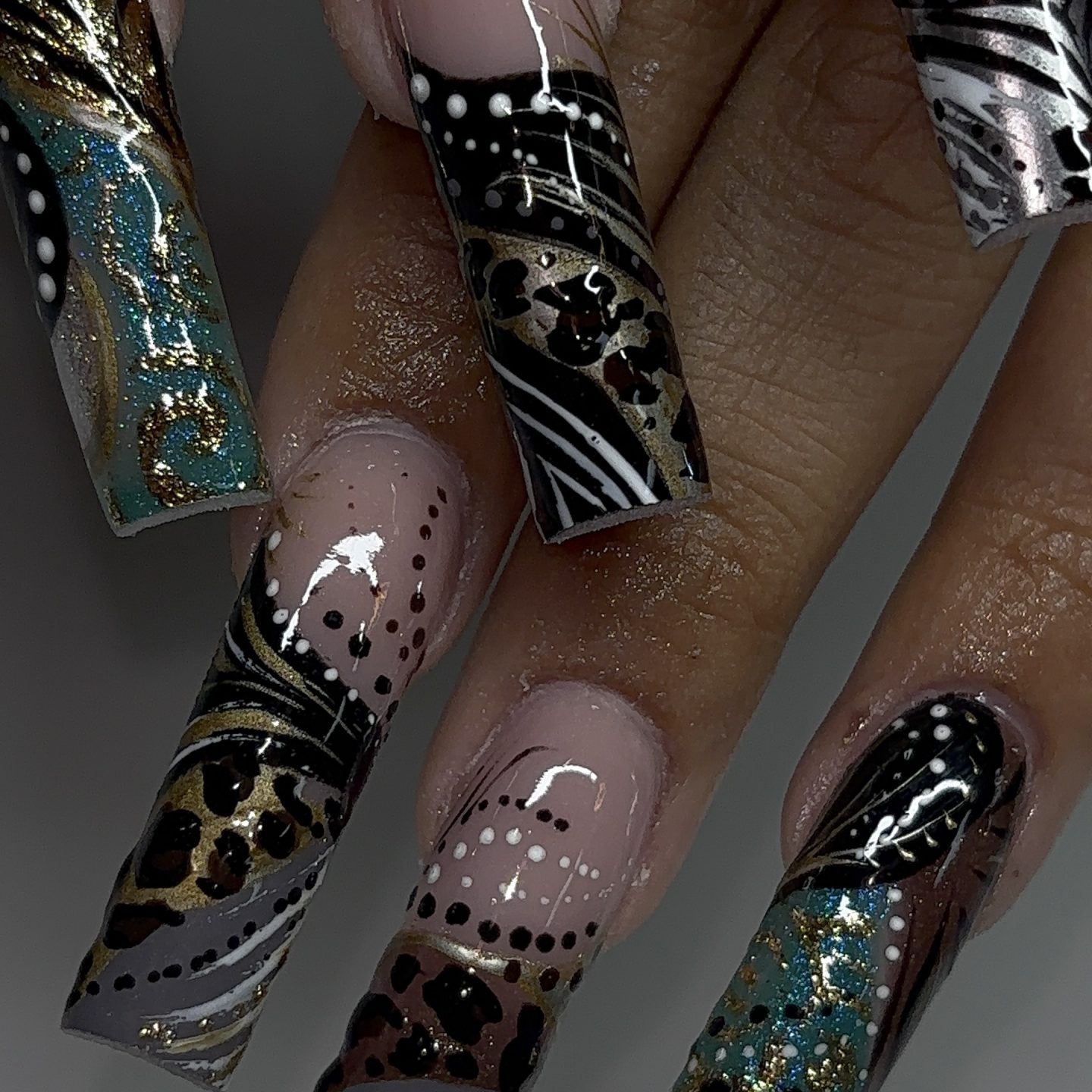 Hand with long, square acrylic nails featuring a mix of animal print, gold glitter, teal accents, and geometric black and white patterns on a nude base.