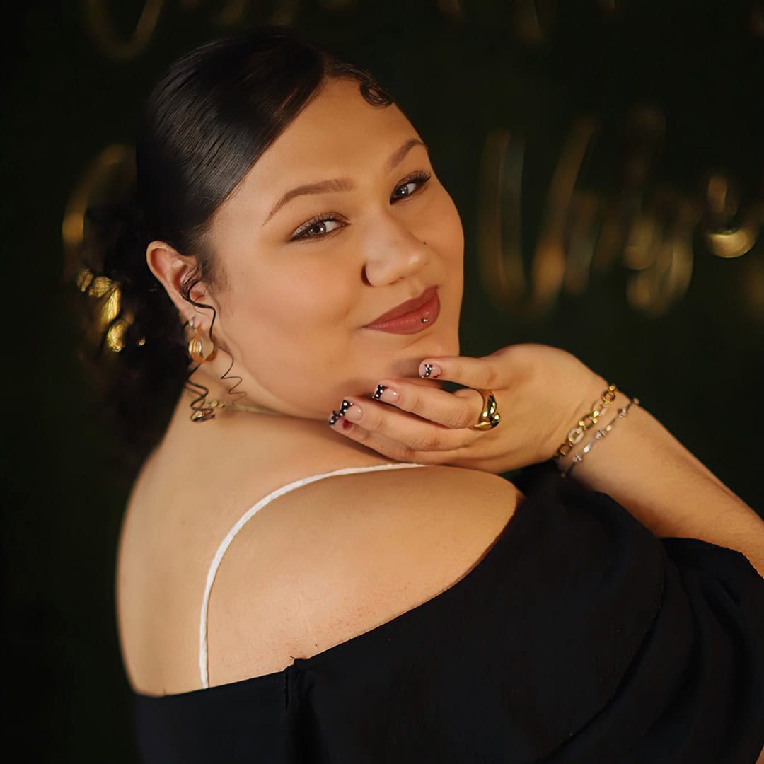 Woman with styled hair and wavy earrings smiles at the camera, hand resting on her chin, wearing an off-shoulder black top and gold jewelry with a dark, blurred background.