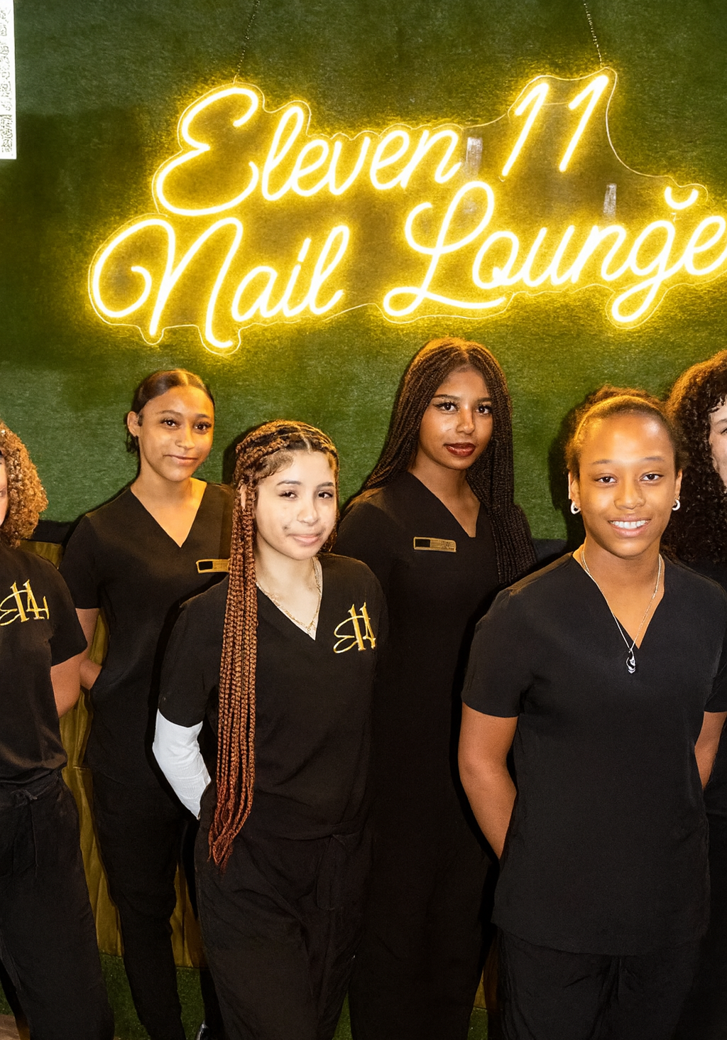 Five women in black uniforms stand in front of a green wall with a yellow neon sign that reads "Eleven 11 Nail Lounge.