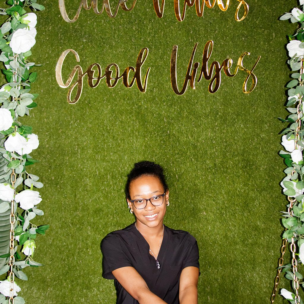 A woman in black scrubs sits on a green bench in front of a green wall with gold text reading "Cute Nails Good Vibes," decorated with white flowers and greenery.
