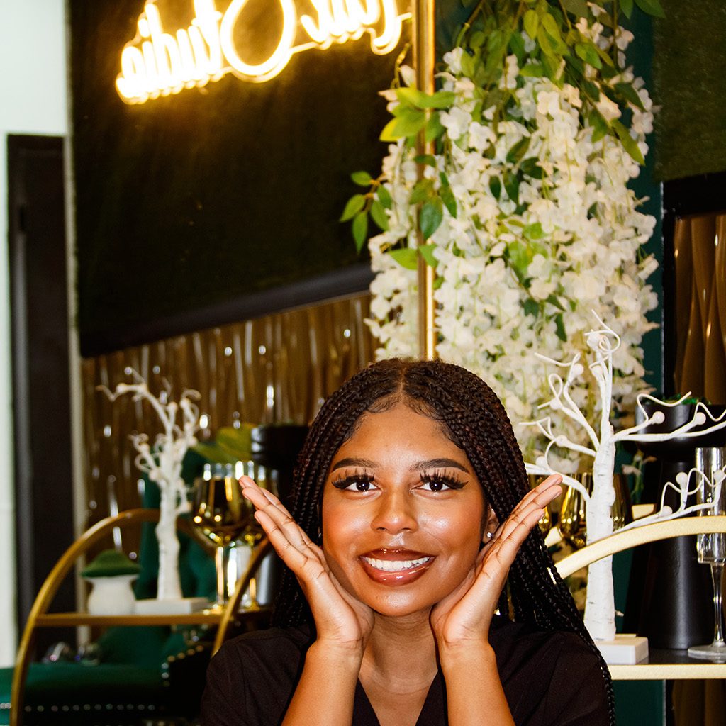 A woman with braided hair and a black top smiles with her hands framing her face in a nail studio, with a neon "Noven 17 Nails Studio" sign in the background.