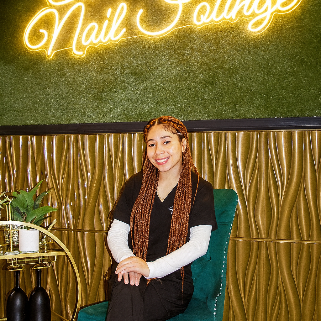 A woman with braided hair sits on a green chair, smiling, in front of a wall with a neon "Nail Lounge" sign and decorative gold paneling.