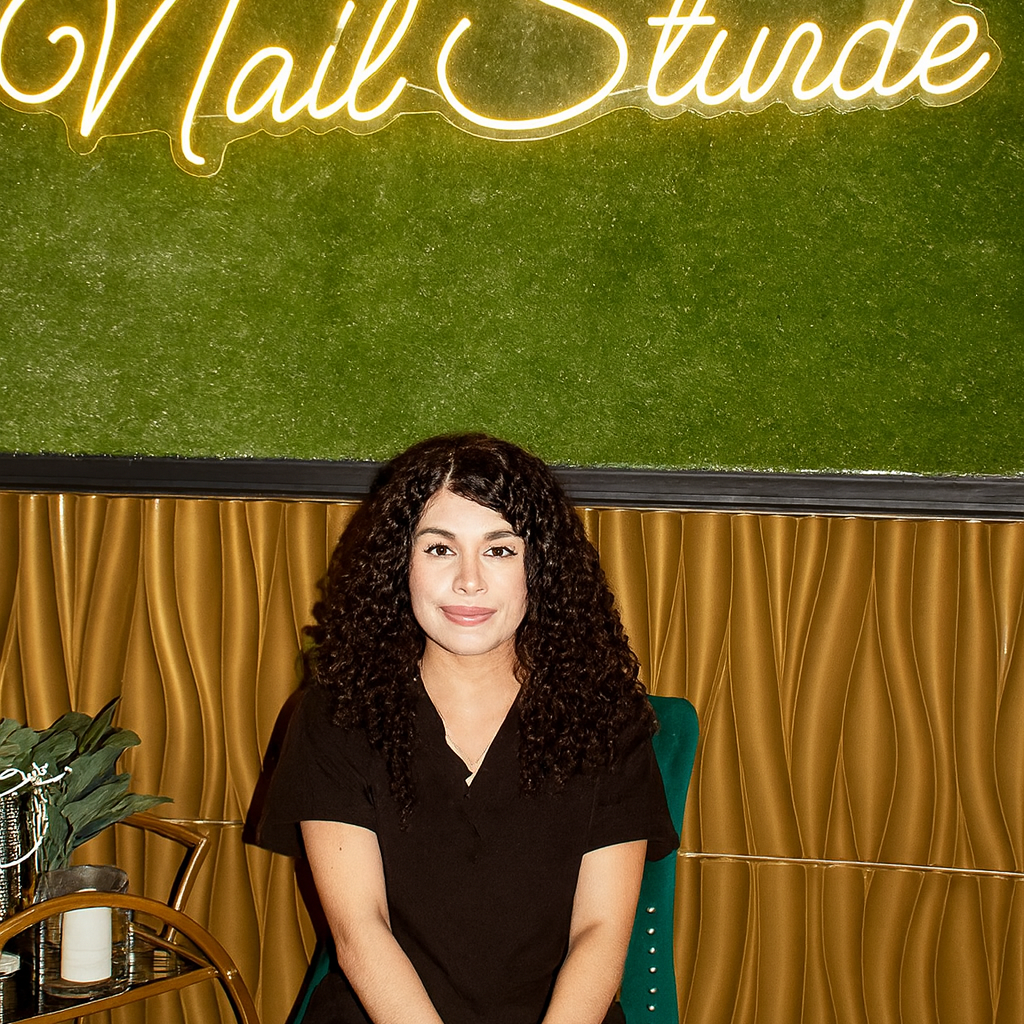 A woman with curly hair sits in a green chair, wearing black scrubs, in front of a neon sign that says "Eleven 11 Nail Studio" on a green and gold wall.